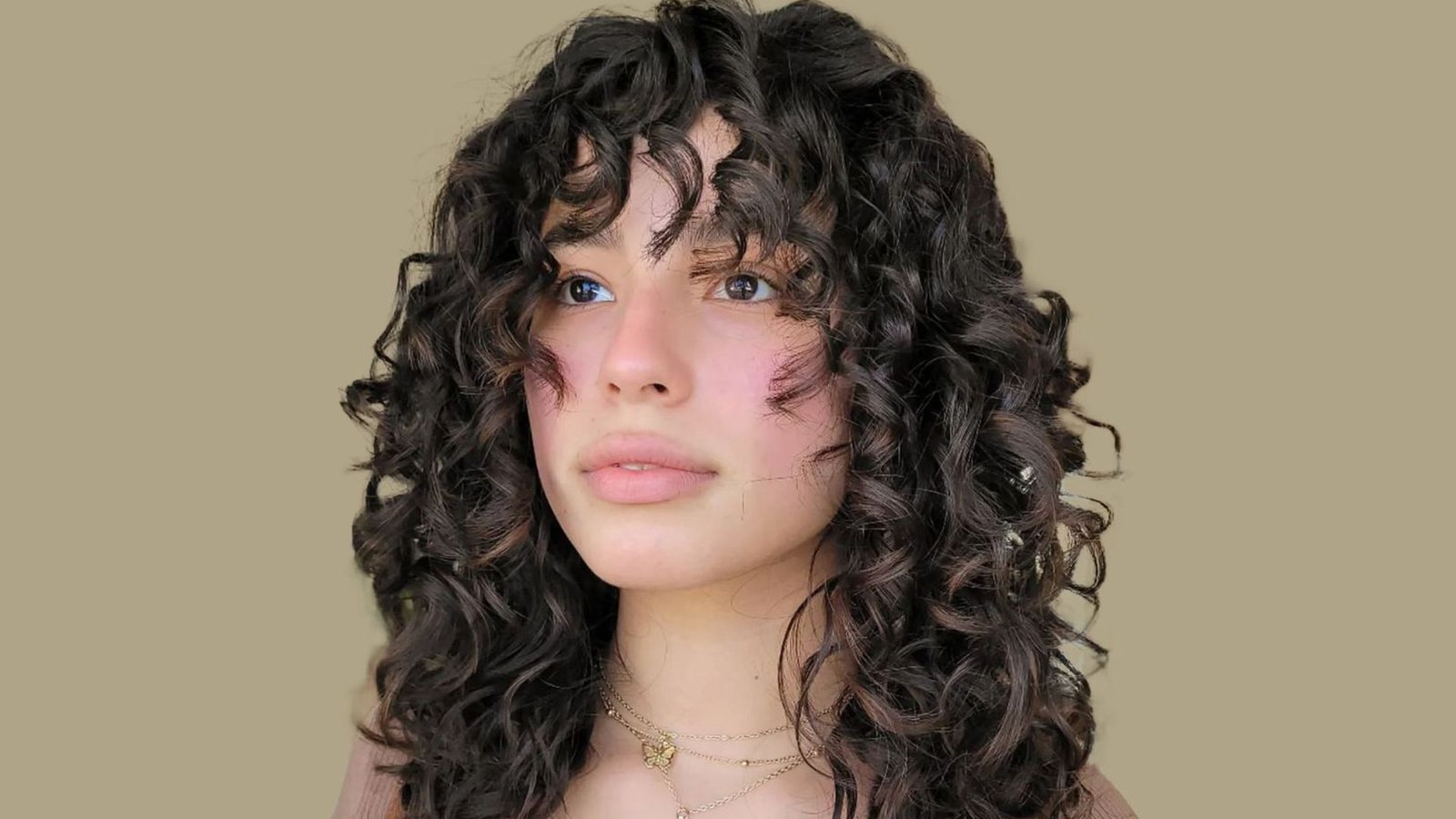 Woman with voluminous curly dark hair and curly bangs. The textured fringe follows the natural curl pattern and sits above the eyebrows.