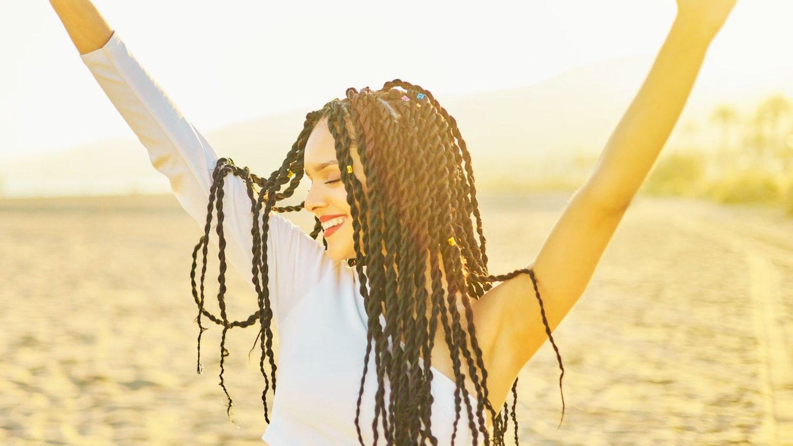 A woman with dreadlocks stands on the beach, showcasing her hairstyle against a backdrop of sand and ocean waves.