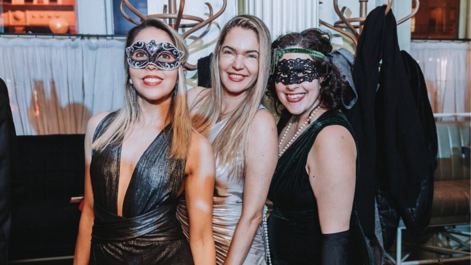 Three women in elegant dresses and masks at a party, smiling warmly. The setting is festive, with coats hanging in the background.
