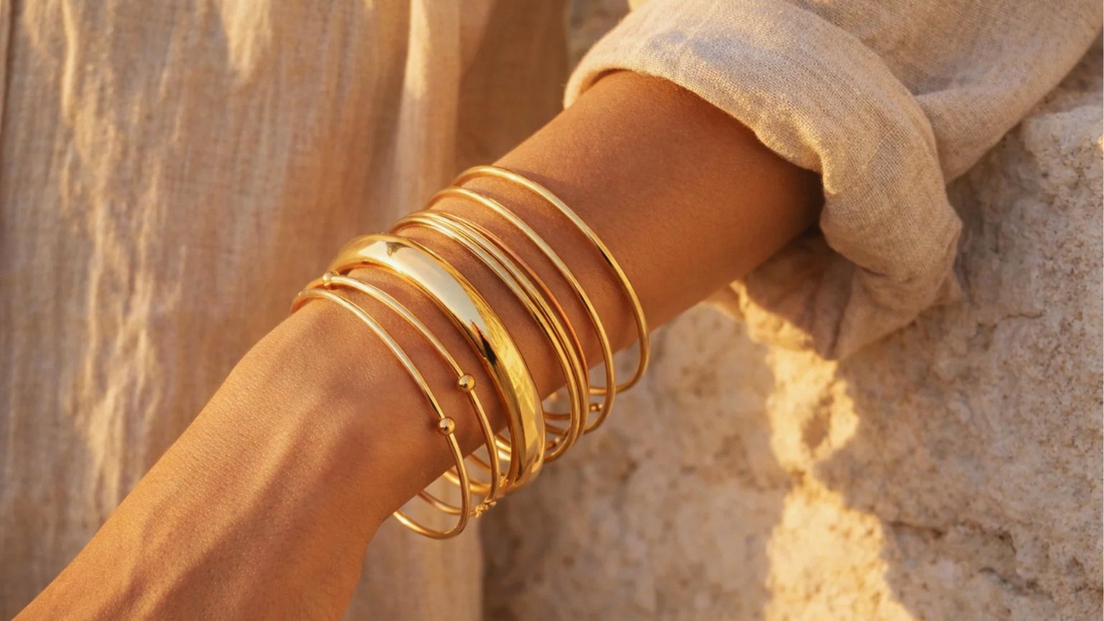 Close-up of a person wearing multiple gold bangles on their wrist, set against a textured sandy background. The tone is warm and elegant.