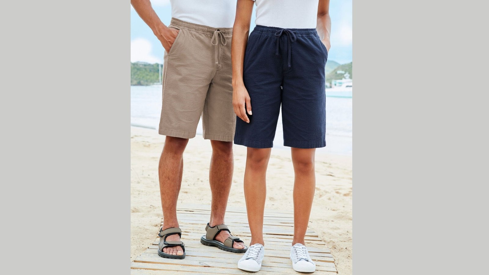 Two individuals on a sandy beach; one wears beige shorts and sandals, the other wears navy shorts and sneakers. A calm sea and sky in the background.