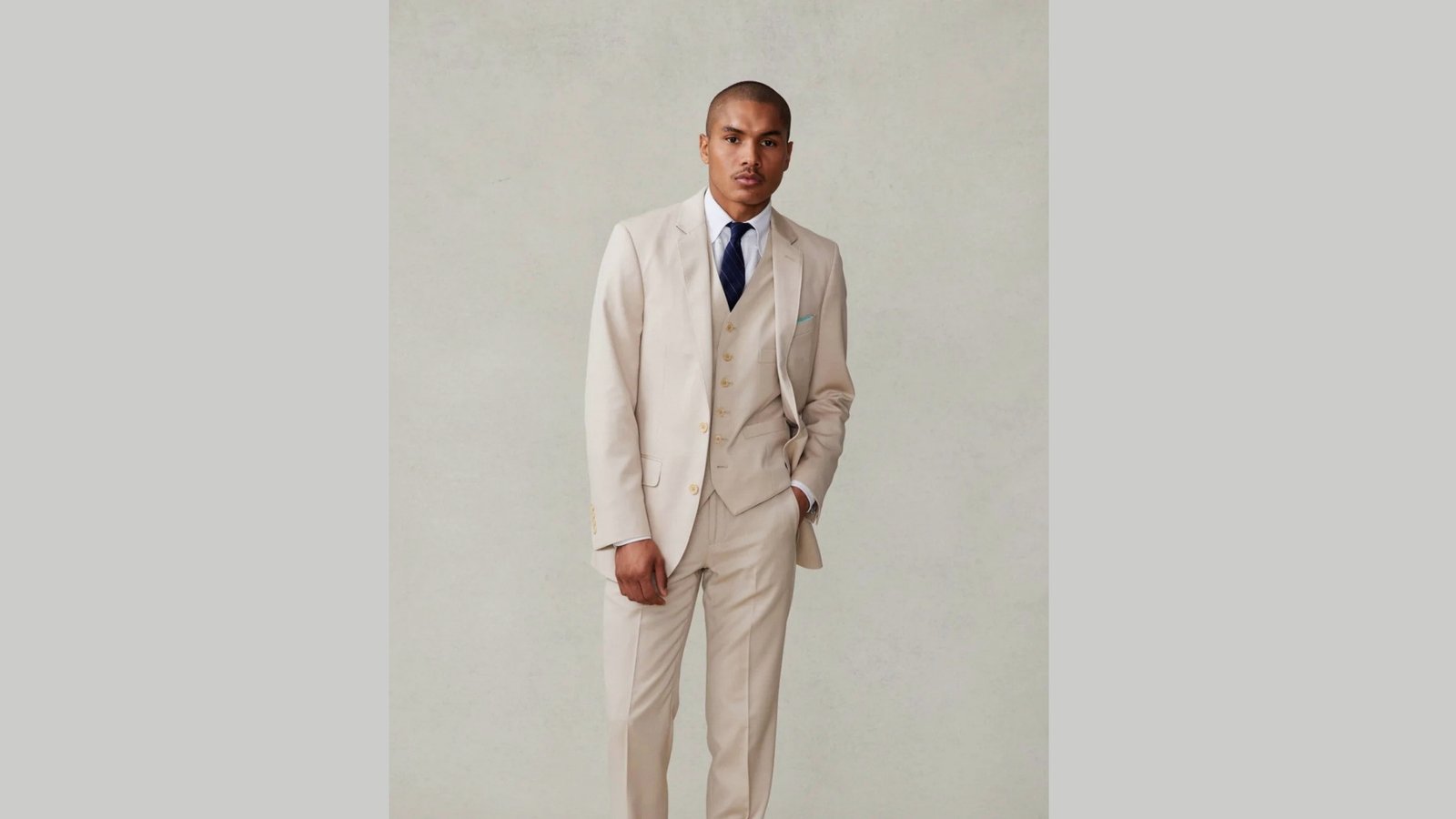 A man in a beige suit and matching vest stands confidently against a neutral background. He wears a blue tie and exudes a formal, composed demeanor.