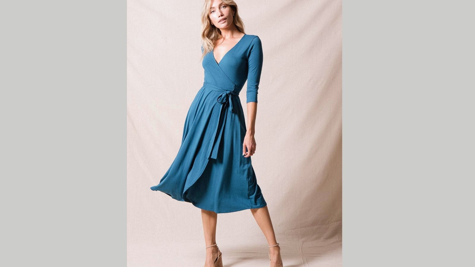 A woman poses gracefully in a blue wrap dress with a V-neck and three-quarter sleeves, exuding elegance. She stands against a beige background.