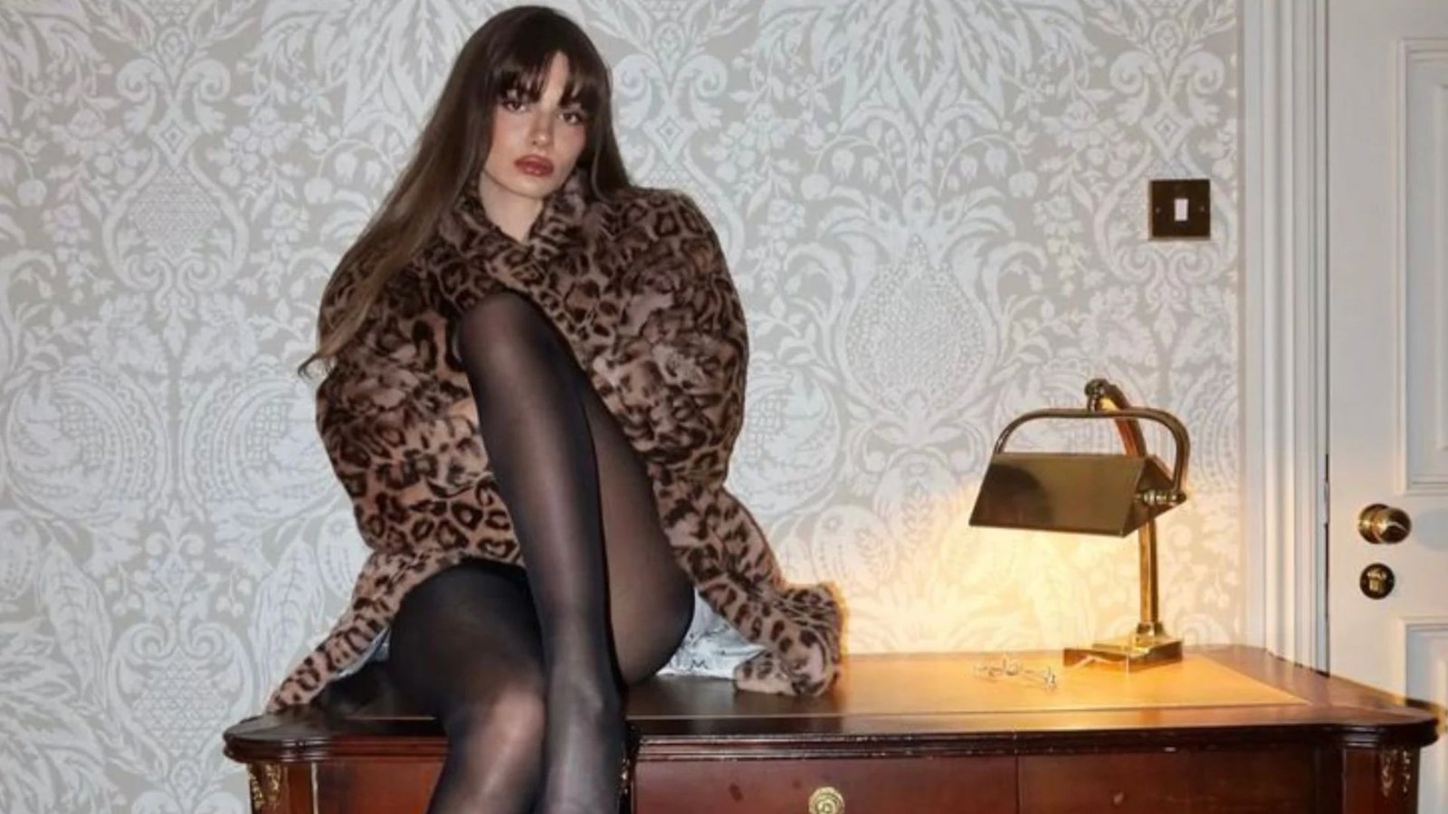 A woman in a leopard-print coat and black tights sits on a wooden desk, with a vintage lamp nearby. The setting has elegant, patterned wallpaper.
