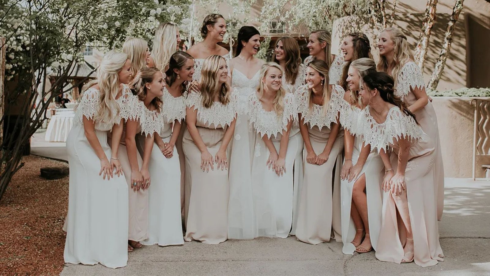 A group of smiling bridesmaids in matching light pink dresses with lace tops pose around a bride in a white gown, set in an outdoor garden.