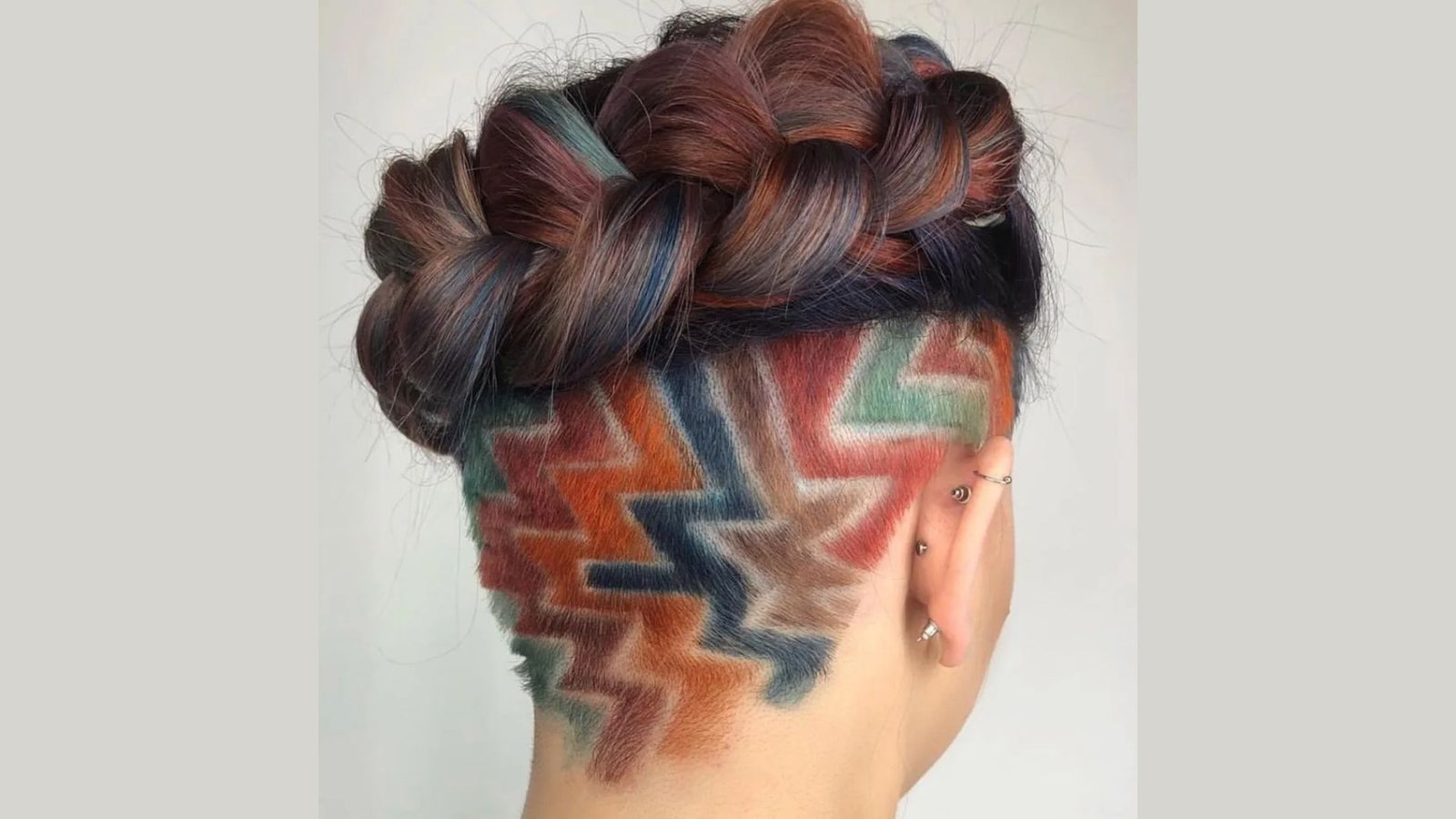 Back view of a woman with a multicolored braided crown revealing a vibrant geometric undercut at the nape. The zigzag pattern features orange, green, blue, and red tones shaved into the buzzed underlayer.
