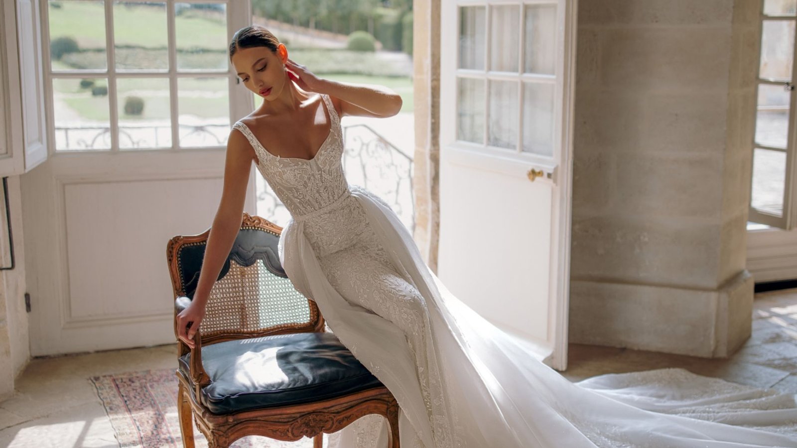 Bride in an elegant, lace wedding dress poses gracefully on a vintage chair by a sunlit window, conveying a serene and romantic atmosphere.