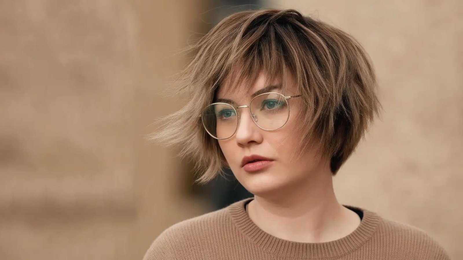 A woman with glasses wearing a brown sweater, featuring a short, edgy shaggy pixie cut.
