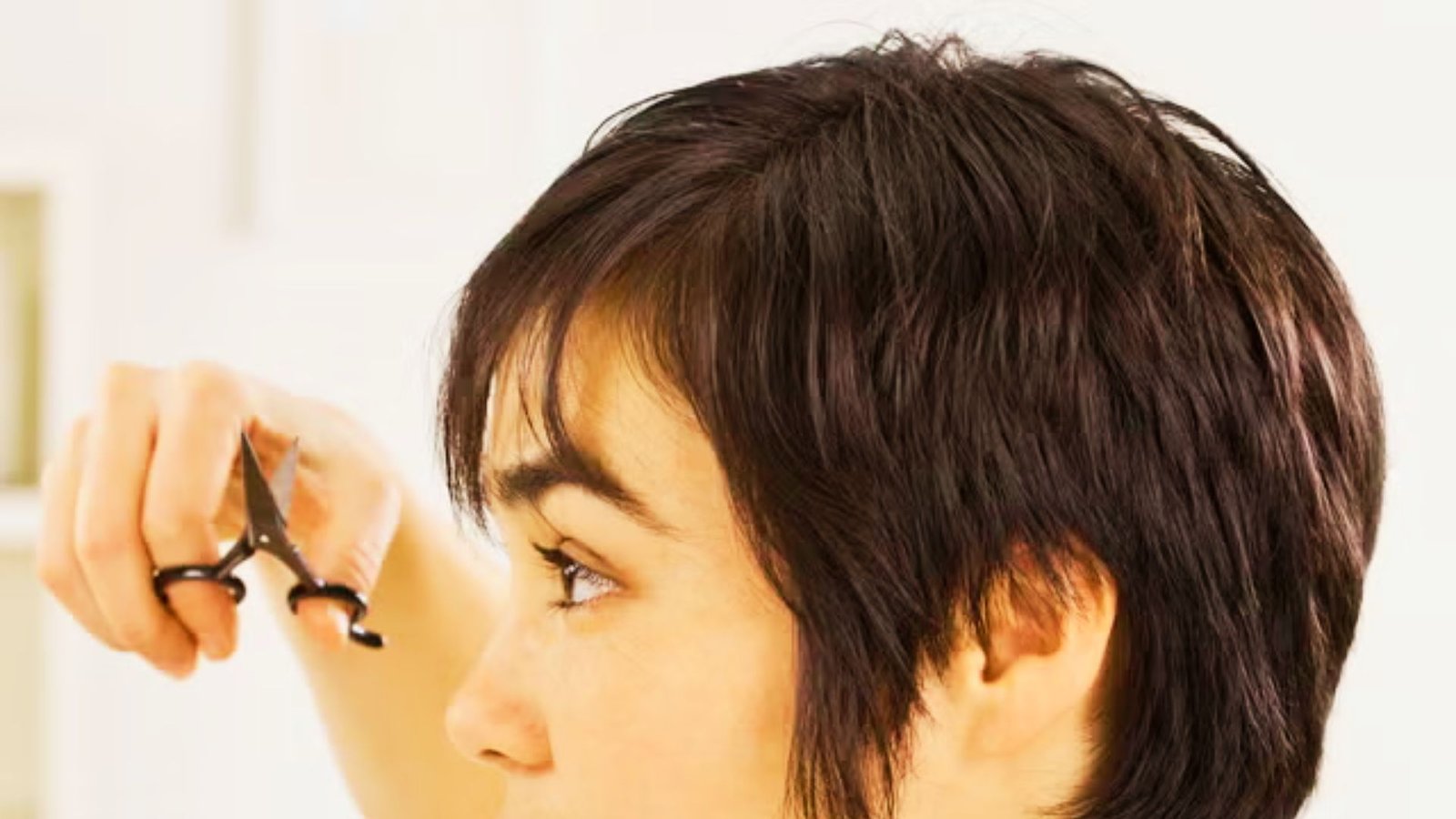 A woman sections her hair with scissors, preparing to cut Bardot bangs at home as part of a step-by-step guide.
