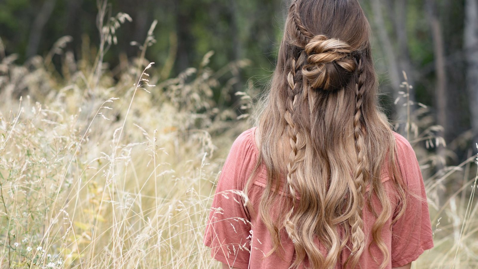 A woman with long hair styled in a half-up bun and bangs stands in a lush green field, enjoying the outdoors.