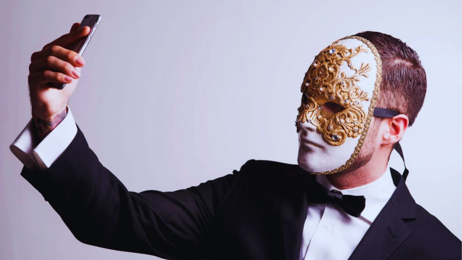 A man in a black tuxedo takes a selfie with a smartphone. He wears an ornate, gold-accented masquerade mask, conveying a mysterious, elegant tone.