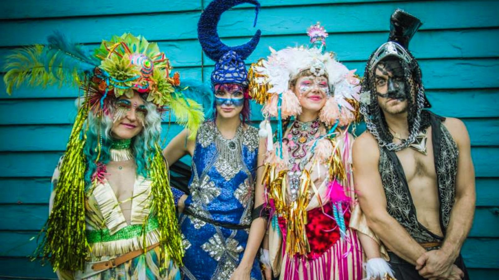 Four people in vibrant, extravagant costumes with colorful feathers and glittery masks stand against a teal wooden wall, conveying a festive and joyful atmosphere.