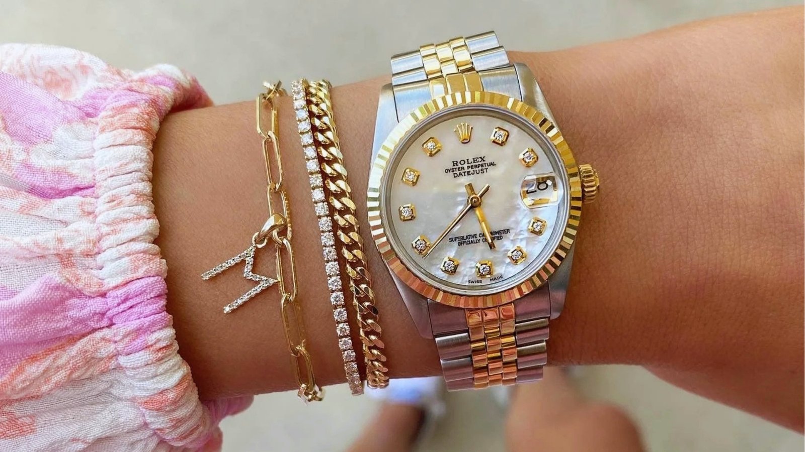 A wrist wearing a Rolex watch with a gold and silver band and diamond markers. The arm also has gold and crystal bracelets, creating a luxurious look.