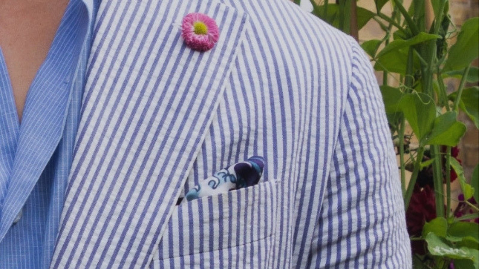 Close-up of a blue and white striped blazer with a pink flower pin and a colorful pocket square. The background features green foliage.