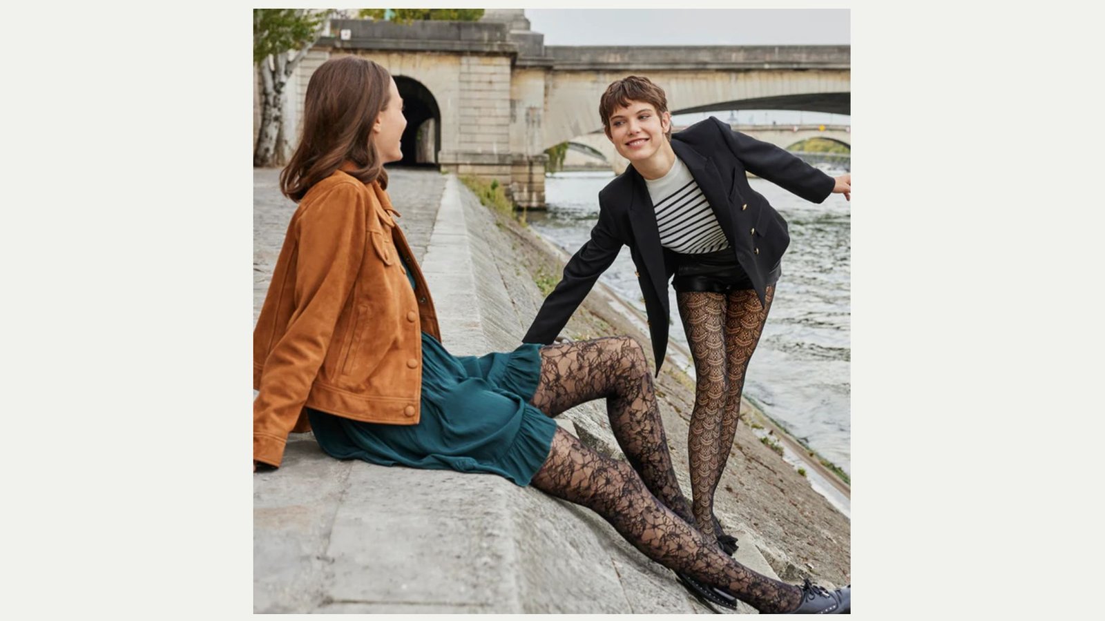 Two women are by a riverside. One, seated in a tan jacket and teal dress, smiles at the other, standing in a striped shirt and black lace tights. Background features a stone bridge, exuding a cheerful, playful vibe.