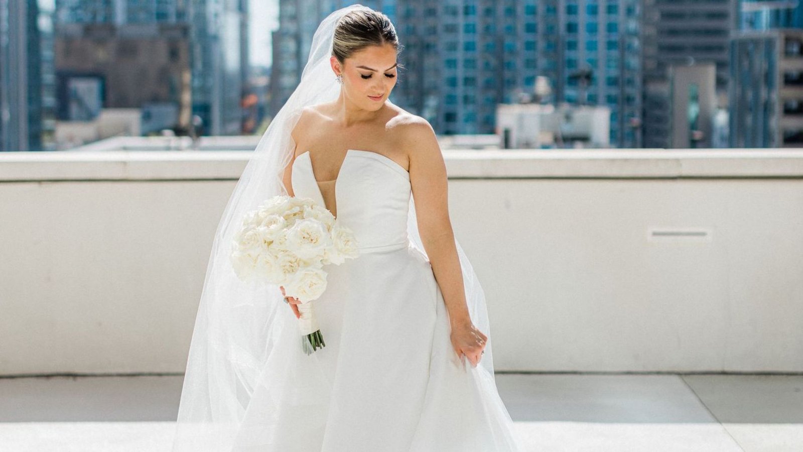 Bride in strapless white gown holds a bouquet of white flowers on a rooftop, with a veil flowing behind her; cityscape backdrop.