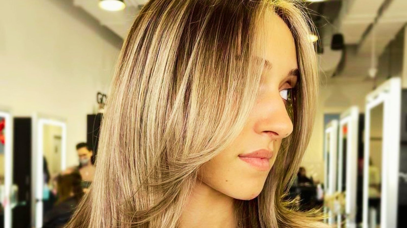 A woman with long blonde hair receiving a haircut in a salon, focusing on blending face-framing layers for Bardot bangs.
