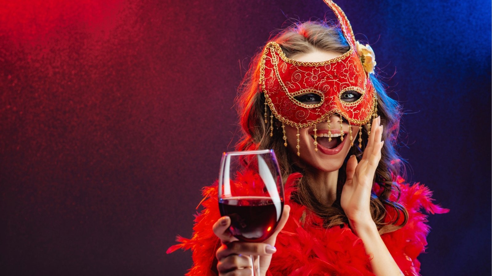 A woman in a red, ornate masquerade mask laughs joyfully, holding a glass of red wine. She wears a matching red feather boa. The mood is festive and vibrant.
