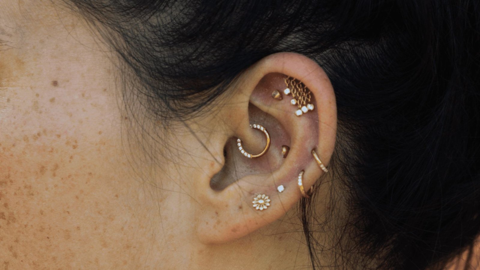 Close-up of an ear adorned with multiple gold and diamond piercings, including a small flower earring. Hair is dark, skin is freckled. Elegant and stylish.