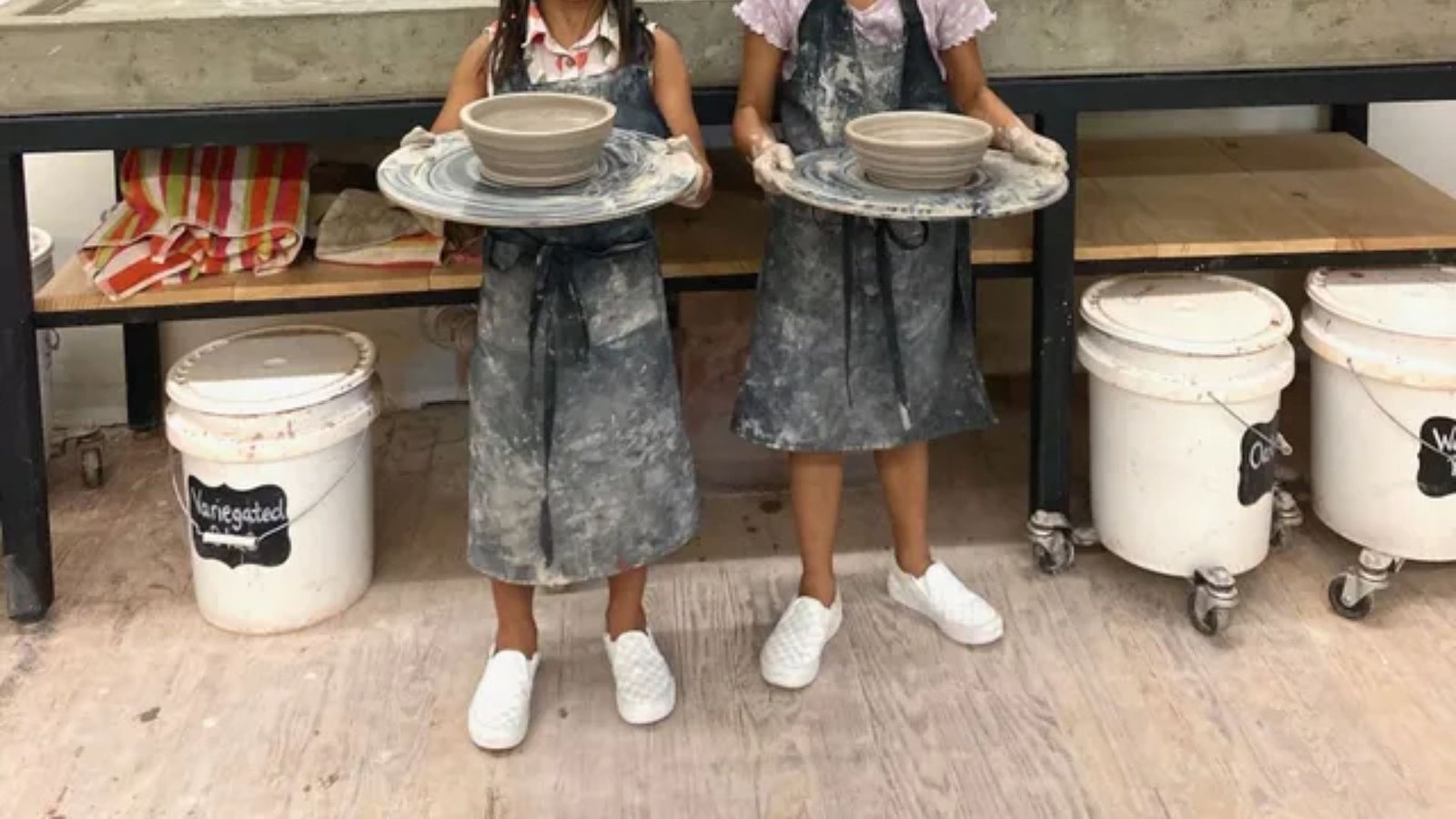 Two children wearing aprons holding clay bowls on pottery wheels. Their clothes are dusty, and they stand in front of a studio workbench with art supplies.