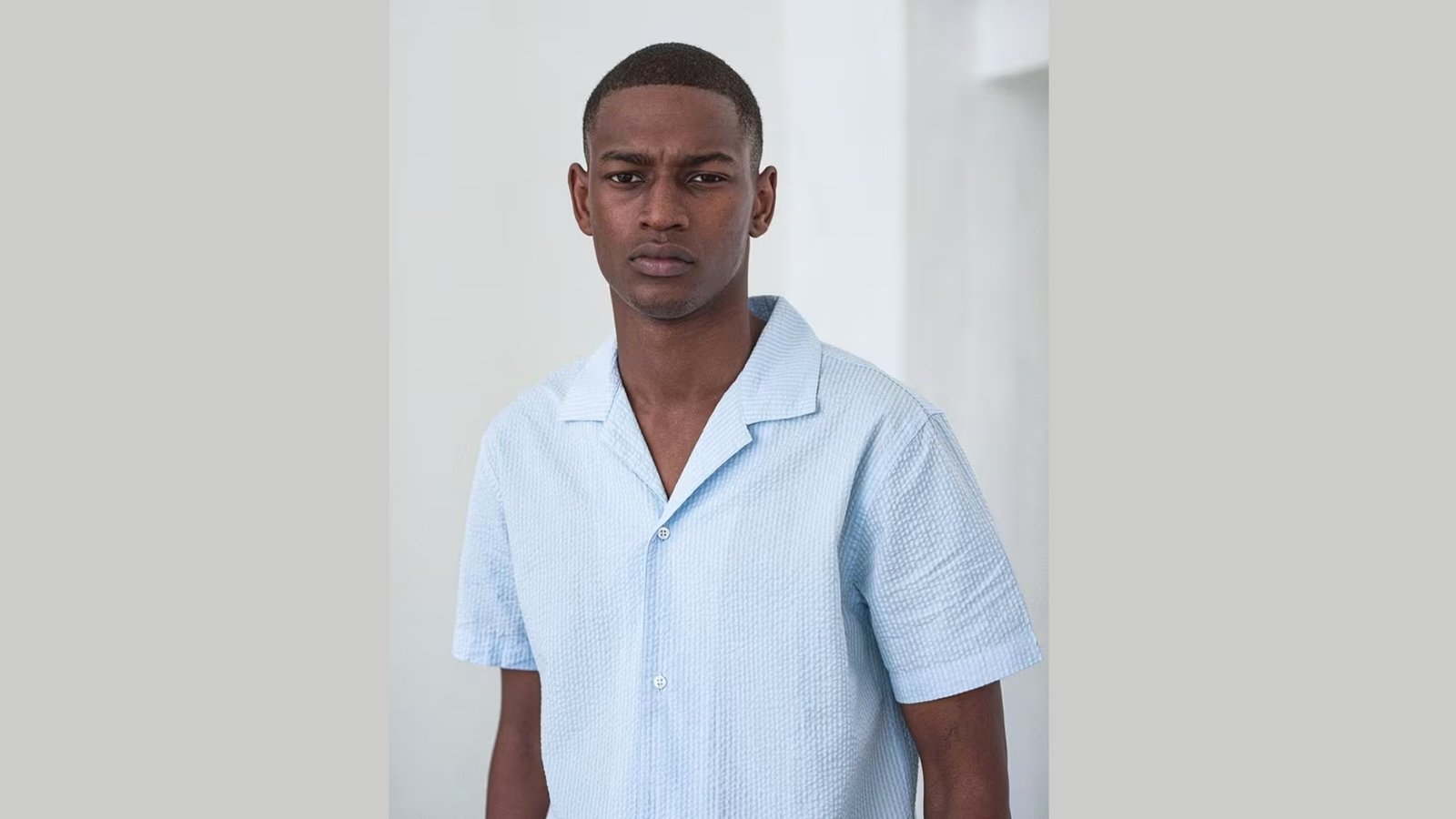 Young man in a light blue shirt stands against a neutral background, looking forward with a neutral expression. The setting is softly lit, conveying calm.