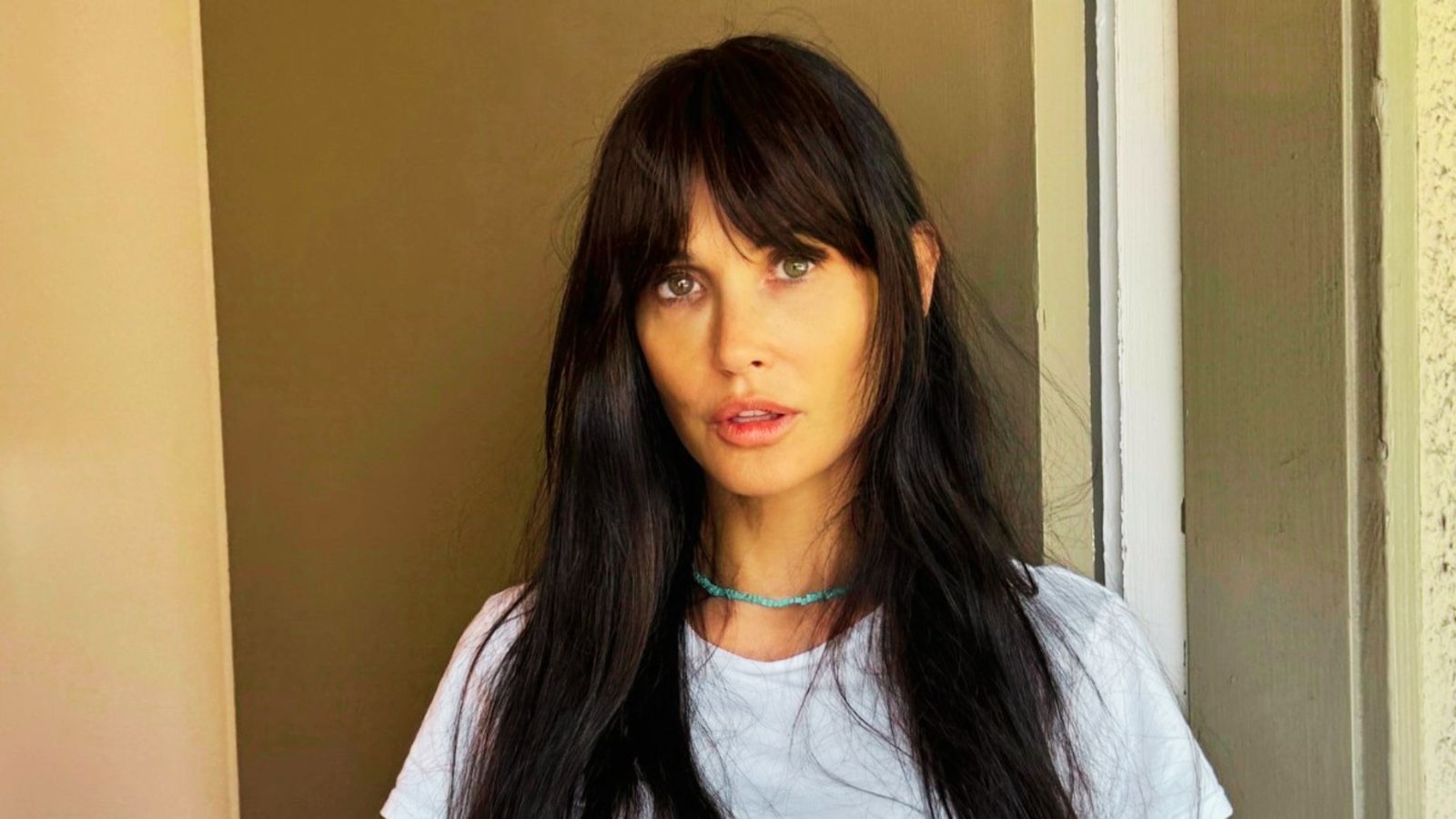 A woman with long black hair wearing a white t-shirt, showcasing a hairstyle known as Bardot bangs.
