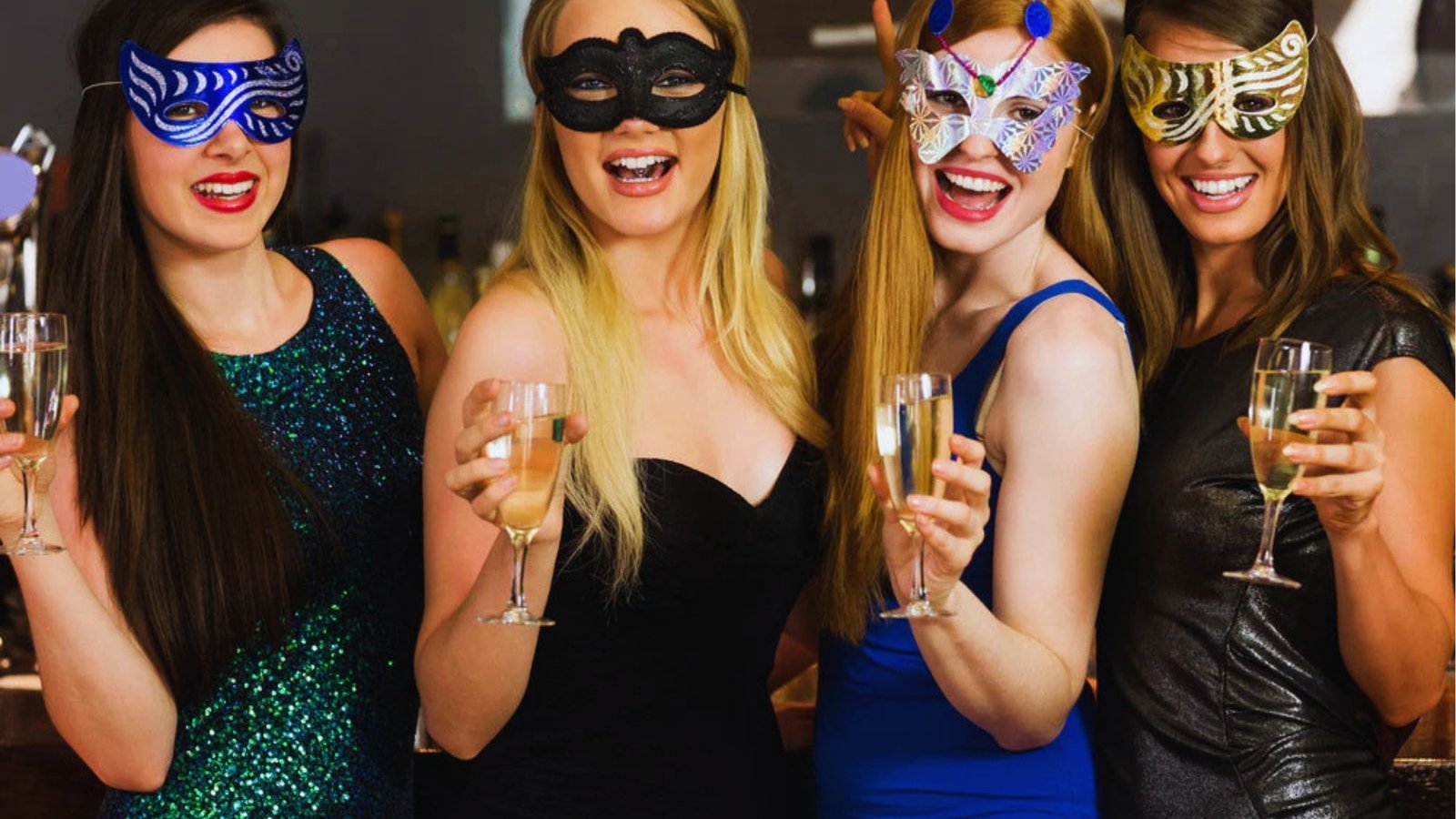 Four women in colorful masquerade masks and elegant dresses hold champagne glasses, smiling with joy. The festive setting suggests a celebratory event.