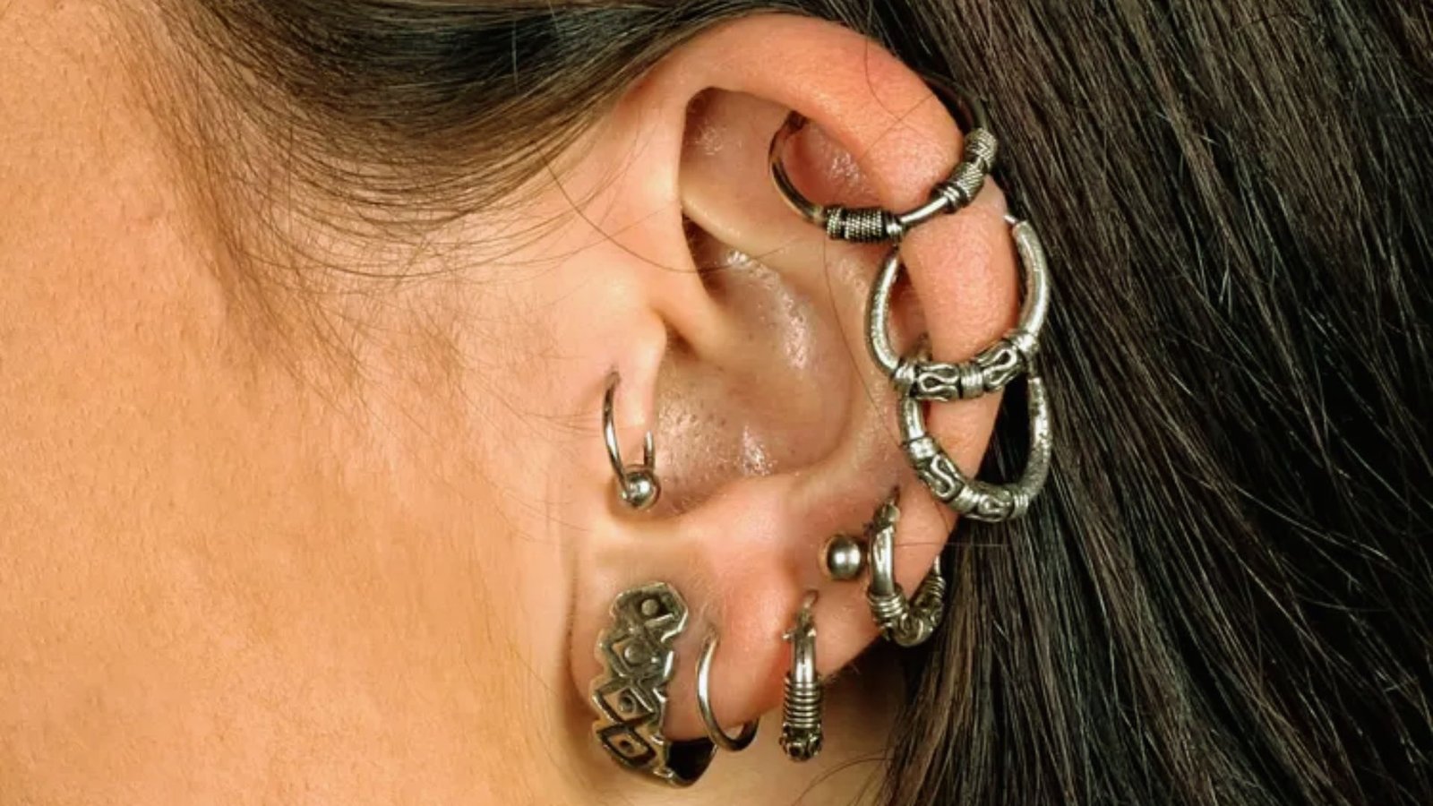 Close-up of a person's ear adorned with multiple silver hoop and stud piercings. The earrings feature intricate designs, creating an edgy tone.