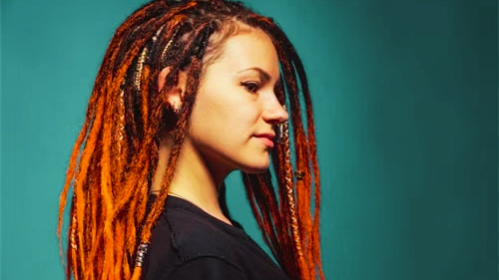 A woman with orange dreadlocks styled in an ombre wicks hairstyle, wearing a black shirt.
