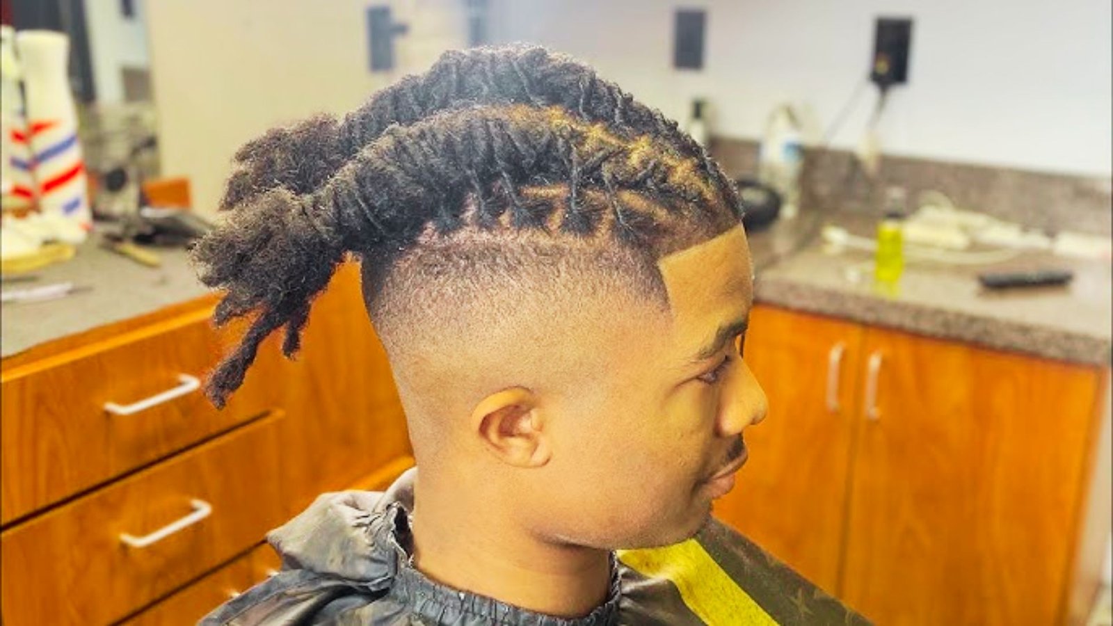 A man with dreadlocks styled in Barrel Twist Wicks sits in a barber shop, surrounded by hair products and tools.