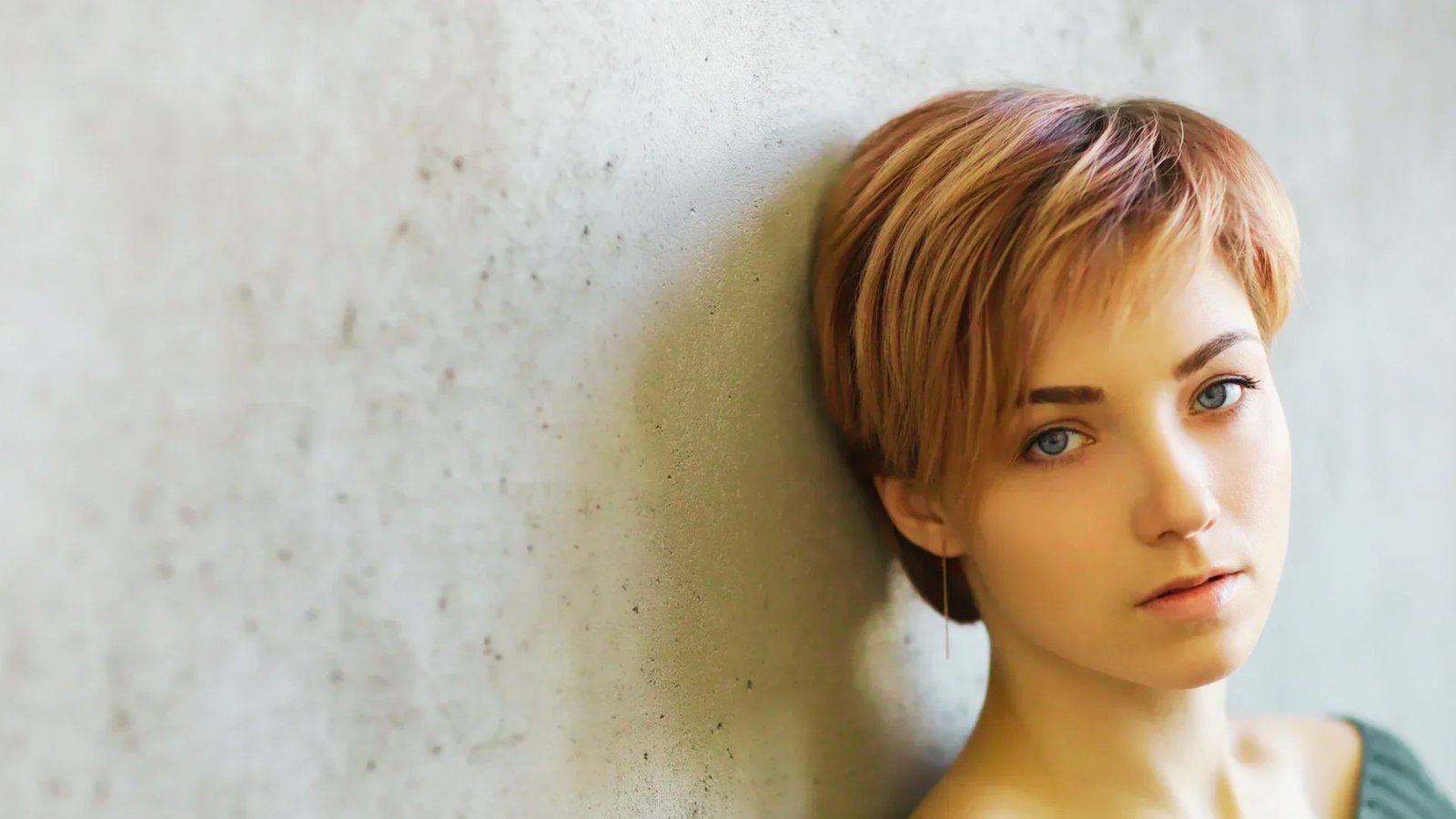 A woman with a tousled fine hair pixie cut leans casually against a wall, exuding a relaxed and confident demeanor.