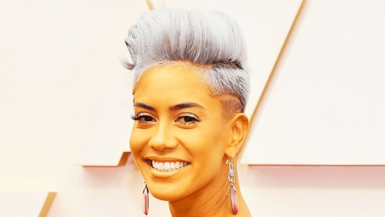 A woman with silver hair styled in a faux hawk pixie cut, wearing earrings and smiling warmly at the camera.